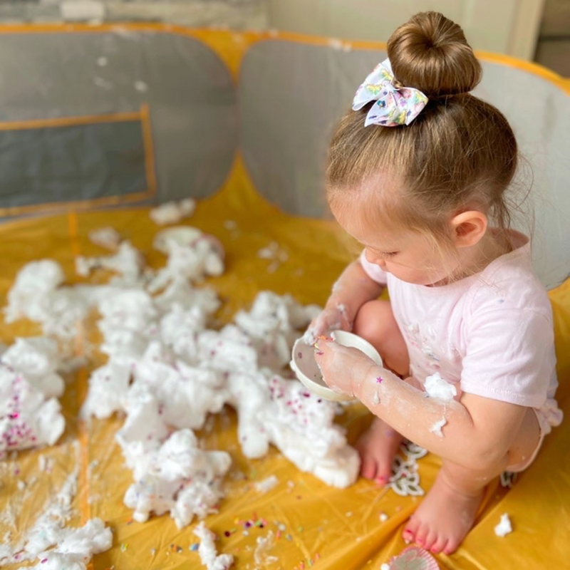 Girl with a bow on her head crouched in a Crafty Pod Playpen whilst playing with shaving foam for messy play