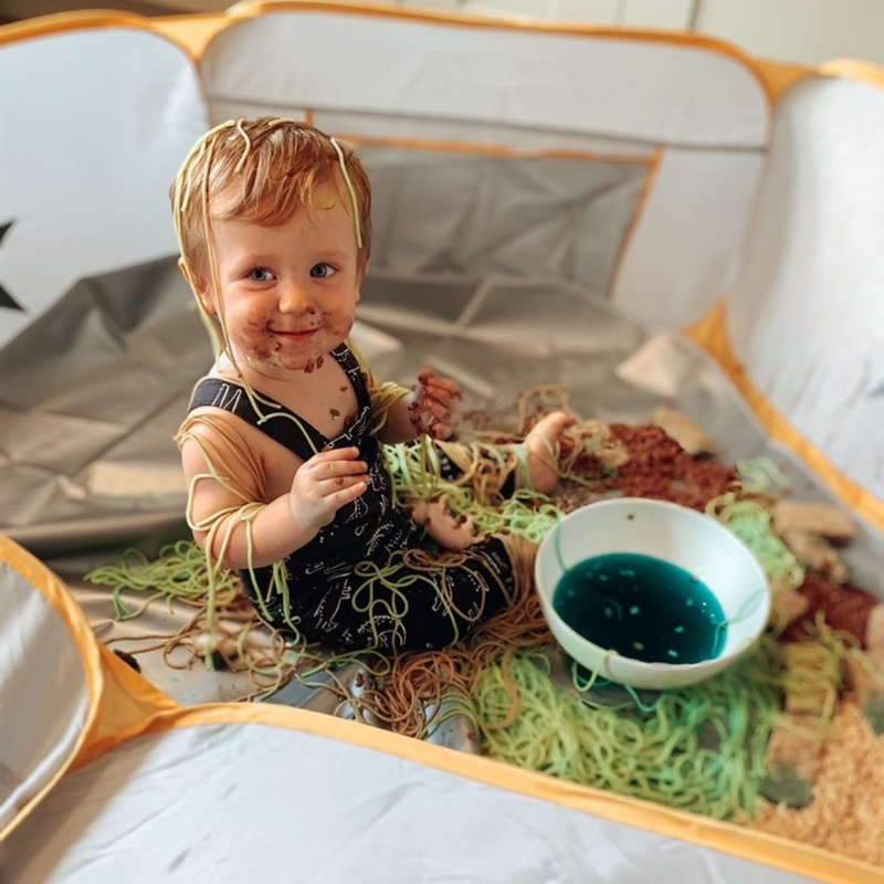 Toddler sat in a Messy Play playpen Crafty Pod with spaghetti on his head, smiling at the camera