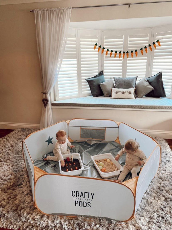 Two toddlers playing in the Crafty Pod fold down playpen set up indoors with a Messy Mat and Sensory Trays